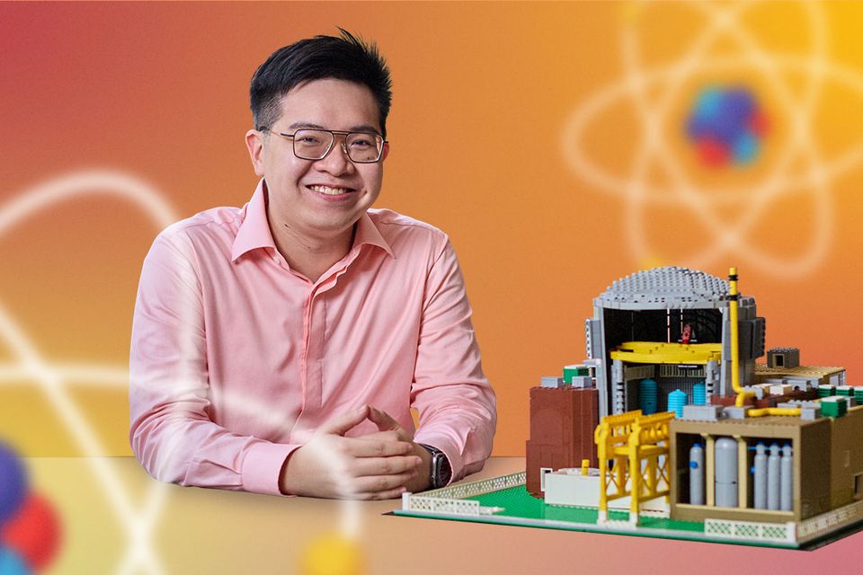 A smiling man beside a brick-built model of a nuclear power plant, on an orange background with atomic graphics